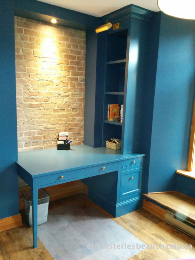 A blue desk with drawers is built into a blue wall with a brick accent. There are shelves with books above the desk and a small lamp on the desk. A window is partially visible on the right.
