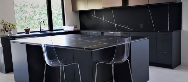 Cuisine moderne avec un îlot central noir, deux chaises transparentes, des armoires sombres et une fenêtre à gauche.