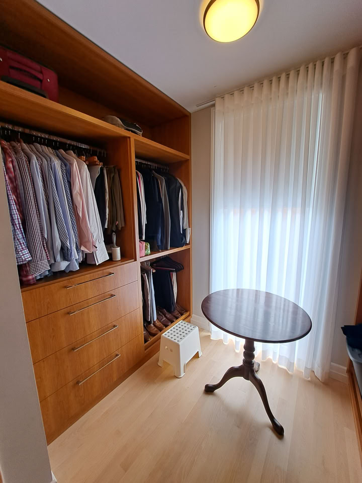 Armoire en bois avec vêtements suspendus, table ronde en bois, rideaux blancs, et lumière au plafond.