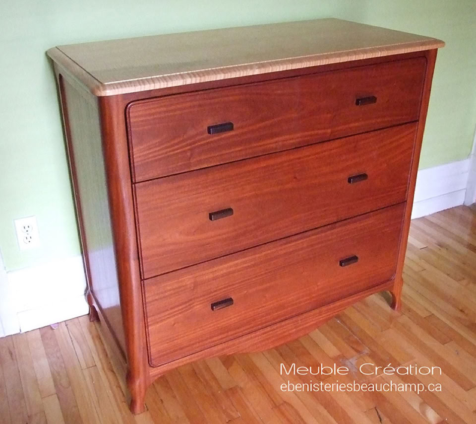 Commode en bois avec trois tiroirs, posée sur un sol en bois clair, contre un mur vert pâle.