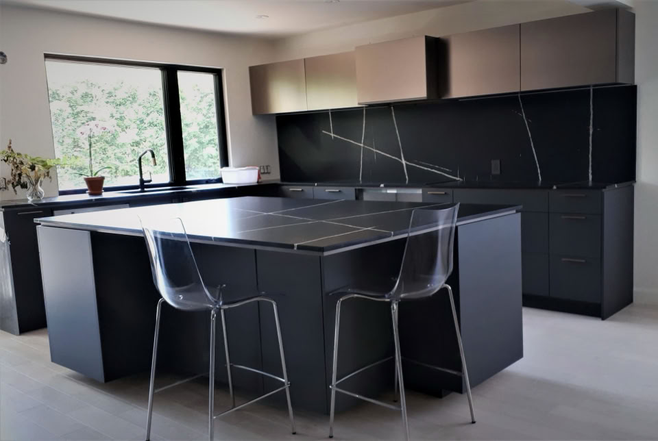 Cuisine moderne avec un îlot central noir, deux chaises transparentes, un évier sous une fenêtre, et des placards sombres.