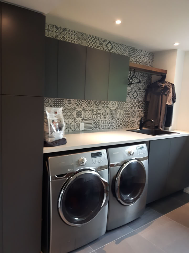 Buanderie moderne avec deux machines à laver argentées, des armoires grises, un plan de travail blanc, et un mur de carreaux à motifs géométriques.