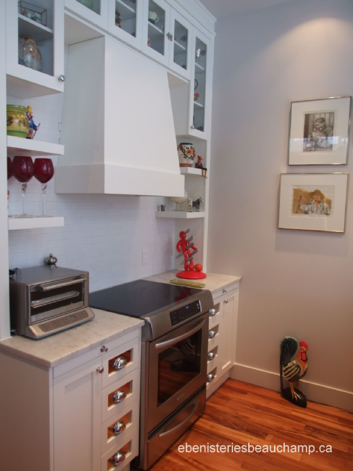 Cuisine moderne avec un four, des placards blancs, des étagères ouvertes avec des verres et des décorations, un sol en bois, et deux tableaux accrochés au mur.