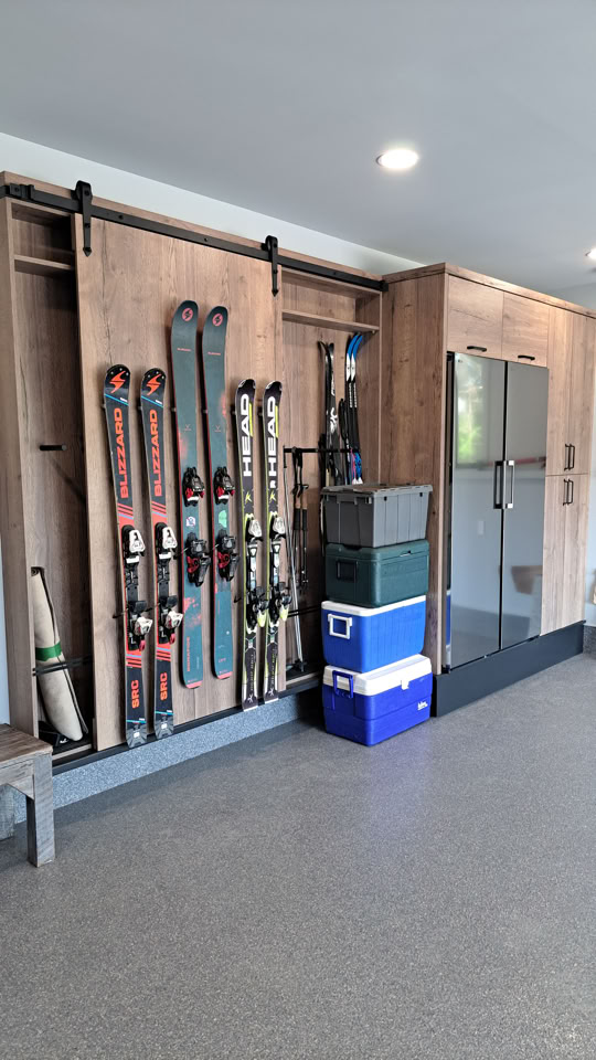 Équipement de ski rangé verticalement dans un garage avec des étagères en bois et des boîtes empilées.