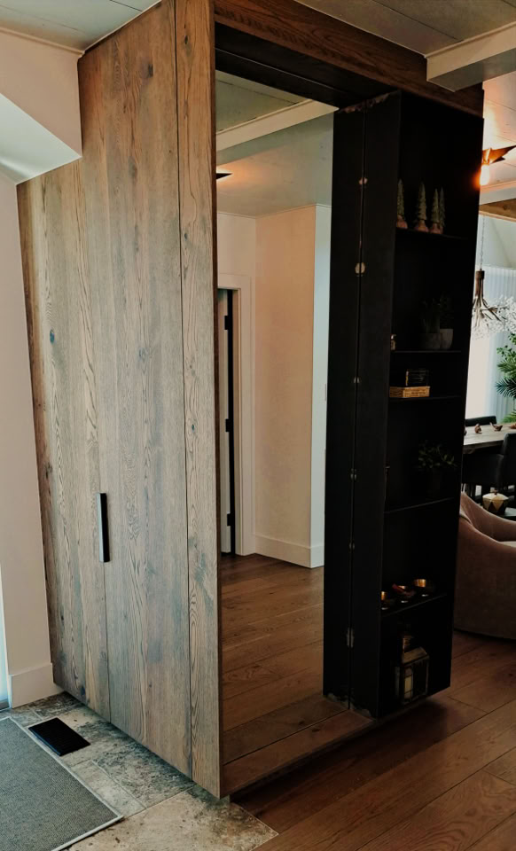 Grande porte en bois avec miroir intégré, ouverte pour révéler des étagères noires à l'intérieur, dans une pièce avec parquet.
