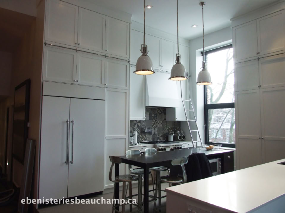 Cuisine moderne avec des armoires blanches, un îlot central avec des tabourets, trois suspensions lumineuses, et une grande fenêtre.