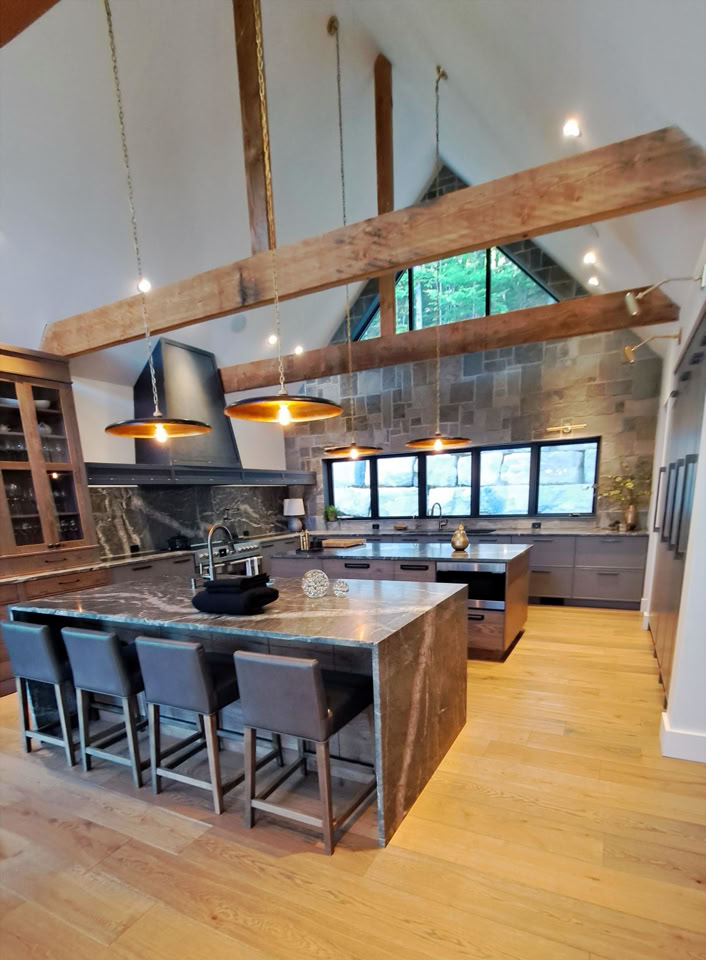 Grande cuisine moderne avec un îlot central en marbre, des tabourets, des poutres en bois apparentes, des luminaires suspendus et des fenêtres donnant sur l'extérieur.