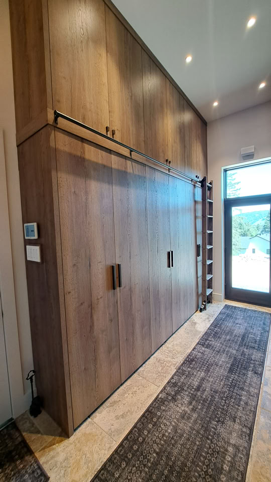 Grand placard en bois avec portes coulissantes, situé dans un couloir avec un tapis au sol et une porte vitrée au fond.