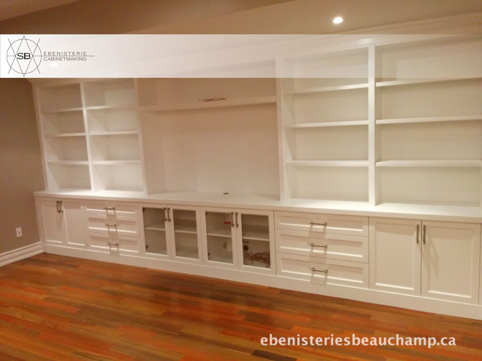 White built-in shelving unit with cabinets and drawers, set against a wall with wooden flooring.