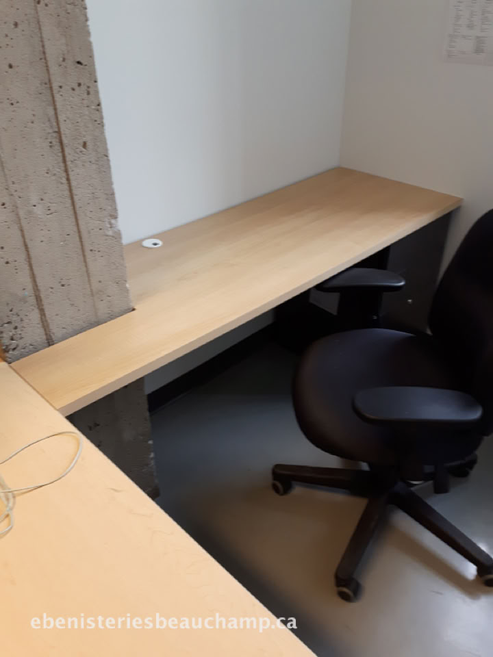 Bureau en bois clair avec une chaise de bureau noire à roulettes, situé dans un coin de pièce avec un mur en béton apparent.