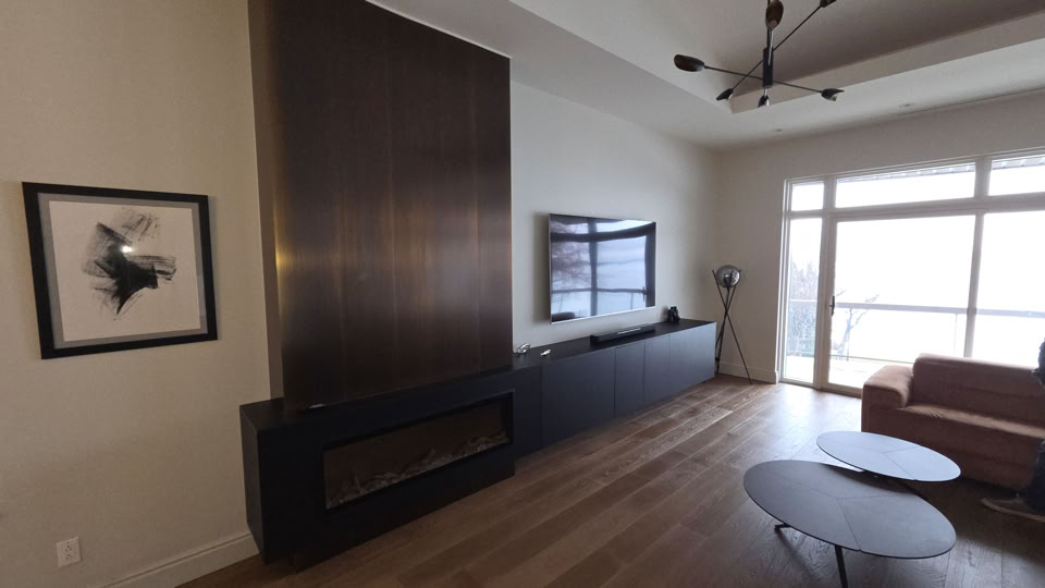 A modern living room with a large dark fireplace, a wall-mounted TV, a sofa, a floor lamp, and two round coffee tables. A framed picture is on the wall, and large windows provide natural light.