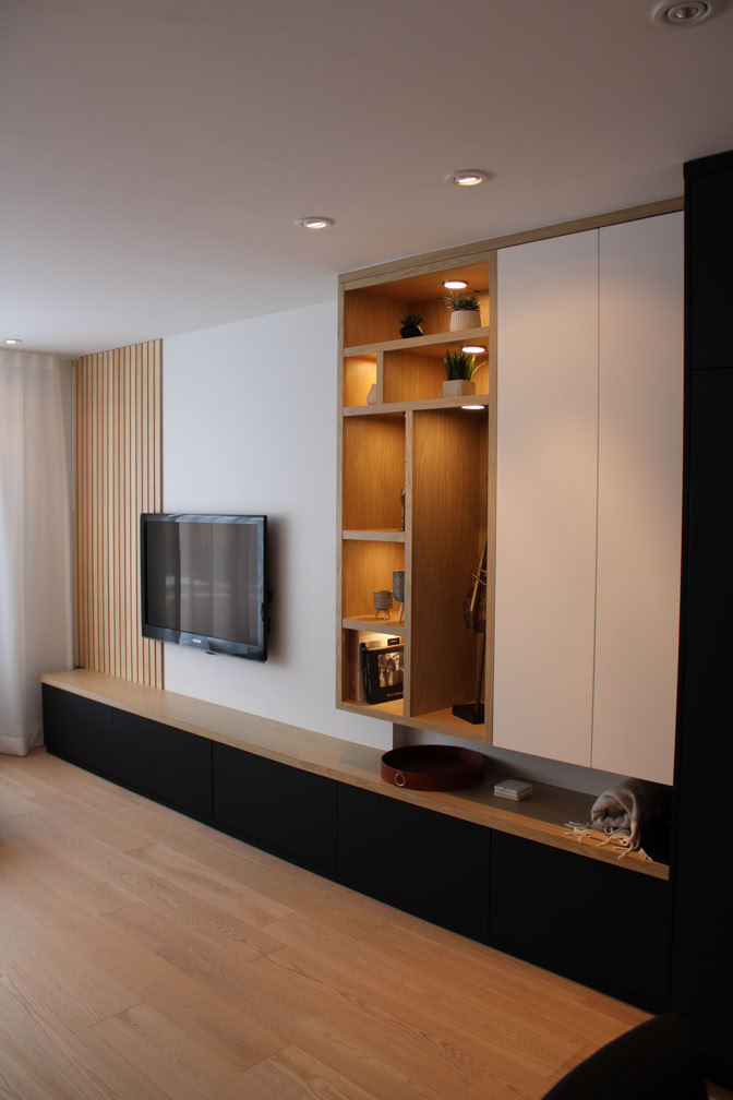 Modern living room with a wall-mounted TV, light wood flooring, built-in shelving with decorative items, and a long black cabinet.