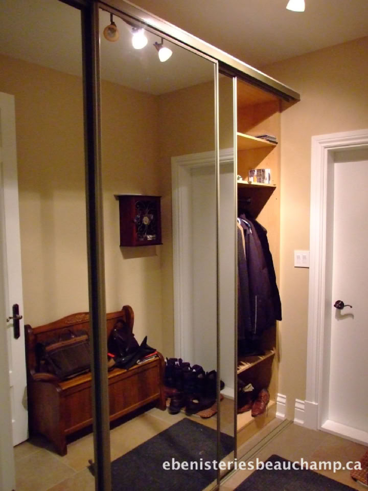 Un couloir avec un placard à portes coulissantes en miroir, un banc en bois, des chaussures au sol, et des manteaux accrochés.