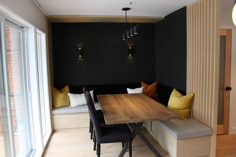 A modern dining area with a wooden table, black chairs, and built-in bench seating with cushions. The walls are dark with two wall sconces, and there is a large window on the left.