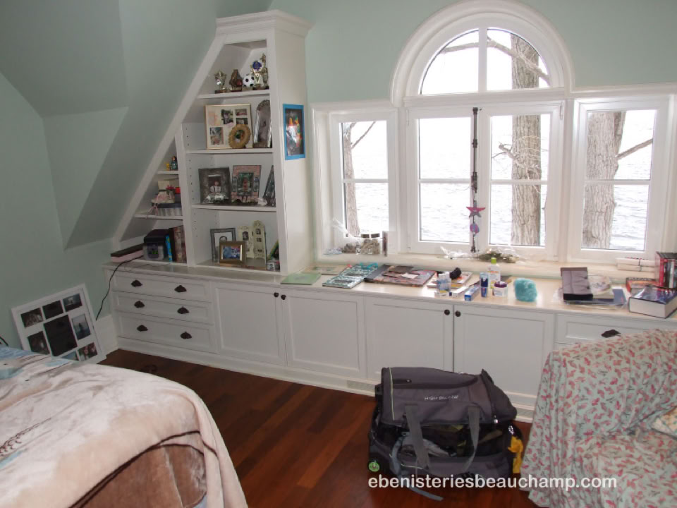 Chambre avec un lit, étagères remplies de livres et objets, fenêtre en arc donnant sur des arbres, et un sac noir sur le sol en bois.