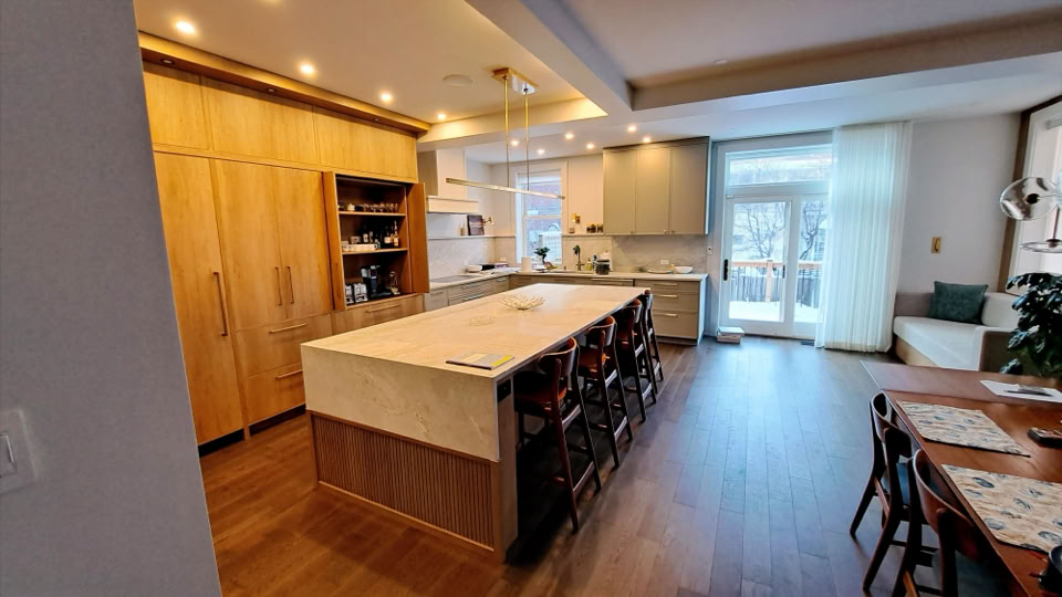 Cuisine moderne avec un îlot central, des armoires en bois clair, un sol en bois foncé, et une table à manger avec des chaises. Fenêtres laissant entrer la lumière naturelle.