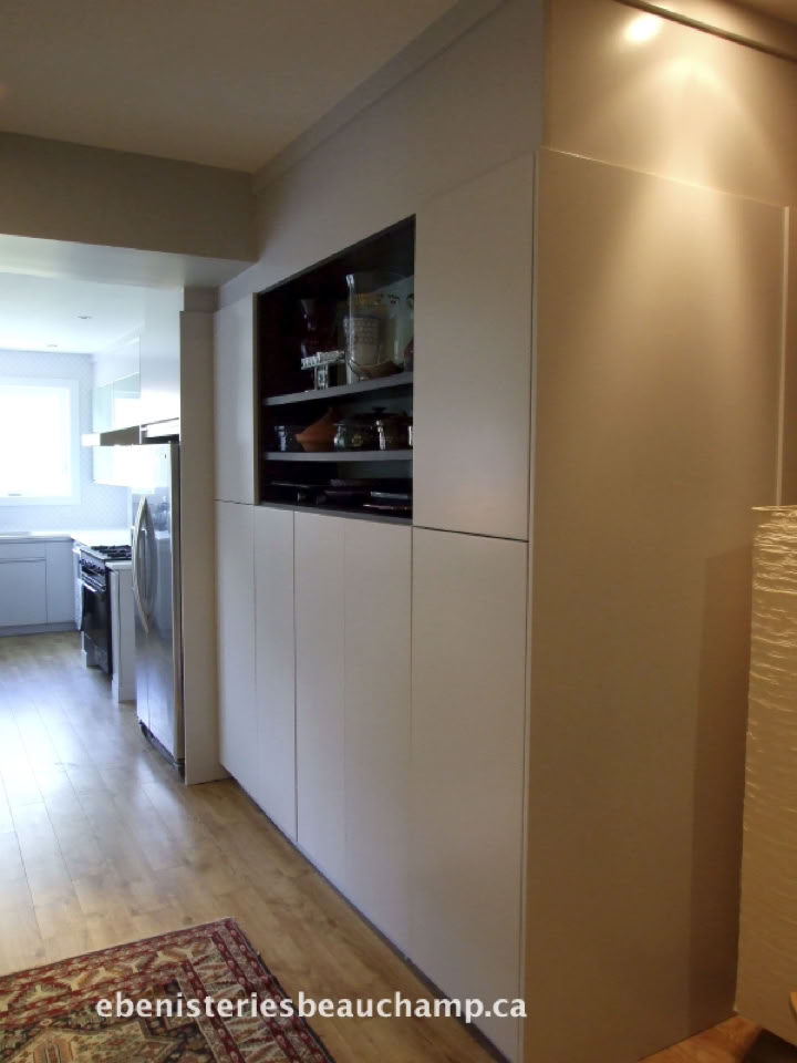 Couloir avec un placard blanc intégré, éclairage au plafond, sol en bois clair, et vue partielle sur une cuisine avec un réfrigérateur en acier inoxydable.