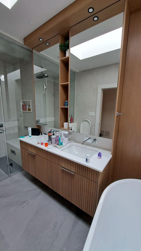 Salle de bain moderne avec un grand miroir, un lavabo encastré dans un meuble en bois, des articles de toilette sur le comptoir, et une douche vitrée à gauche.