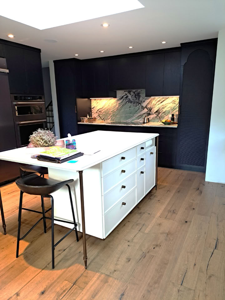 Cuisine moderne avec un îlot central blanc, tabouret noir, plancher en bois, et mur de fond sombre avec un dosseret en marbre.