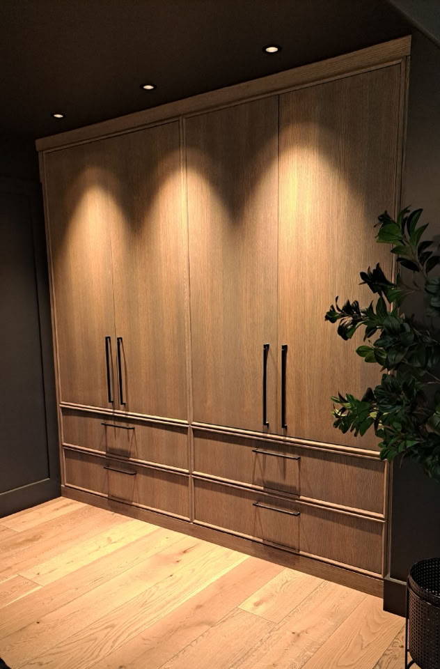 Armoire en bois avec trois portes et poignées métalliques, éclairée par des spots au plafond, sur un sol en bois clair.
