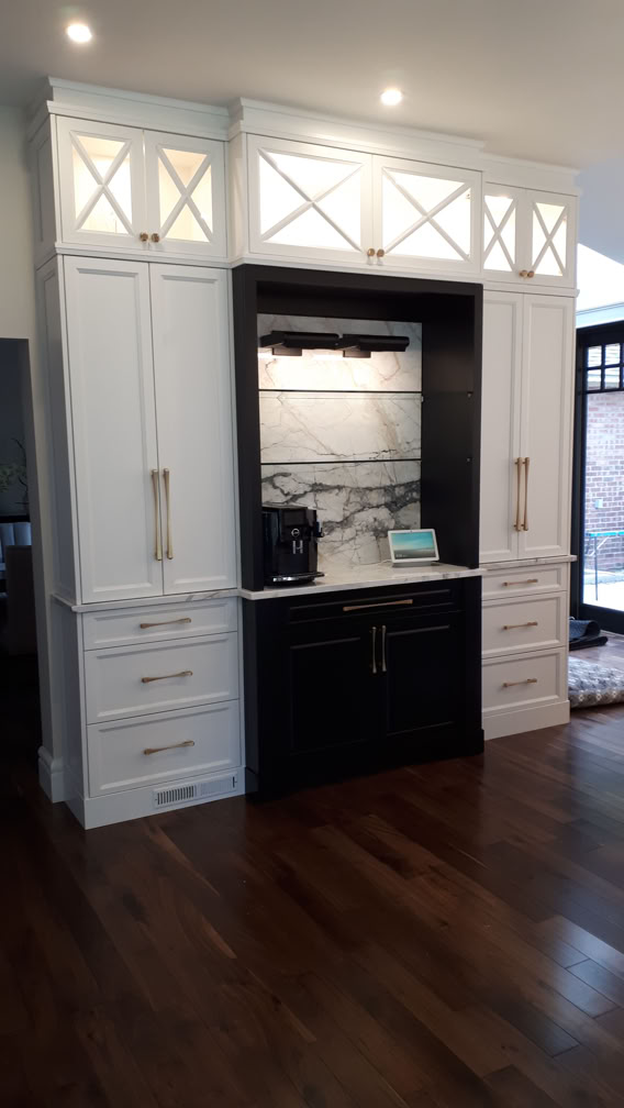 A kitchen area with white cabinets, a black countertop, and a marble backsplash. There are drawers and cabinets on either side, with decorative glass panels above. The floor is wooden.