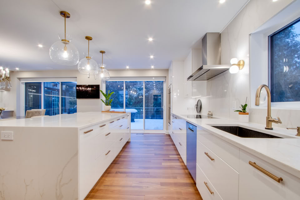 Cuisine moderne avec des armoires blanches, un îlot central, un évier, des luminaires suspendus, un plancher en bois et de grandes fenêtres donnant sur l'extérieur.