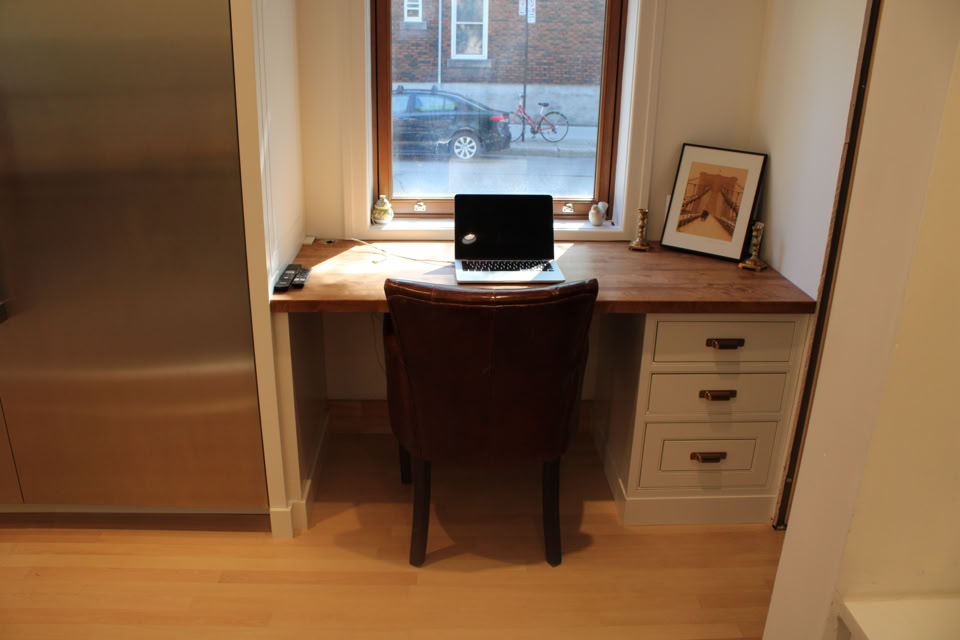 Bureau en bois avec un ordinateur portable, une chaise marron, une fenêtre en arrière-plan, et un cadre photo sur le côté droit.