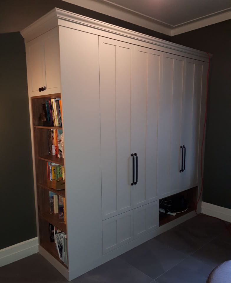 Armoire blanche avec des poignées noires, étagères de livres sur le côté gauche, et une ouverture en bas.