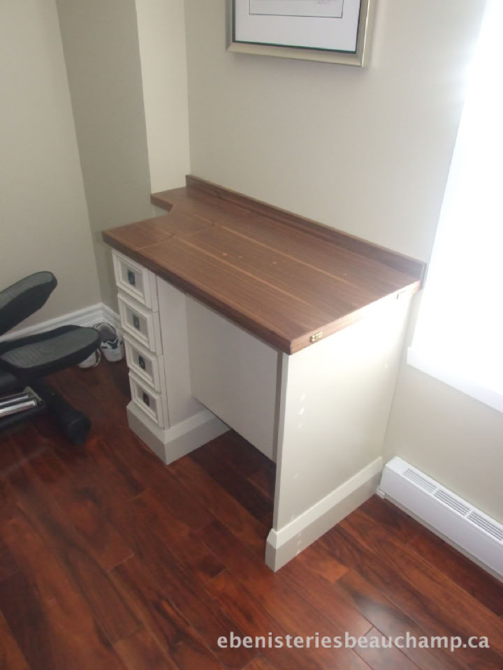 Petit bureau en bois avec trois tiroirs blancs, situé dans un coin de la pièce avec un sol en bois foncé.